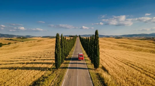 Strada di campagna toscana con colline di grano dorato, cipressi allineati e pullman turistico rosso in lontananza sotto cielo azzurro